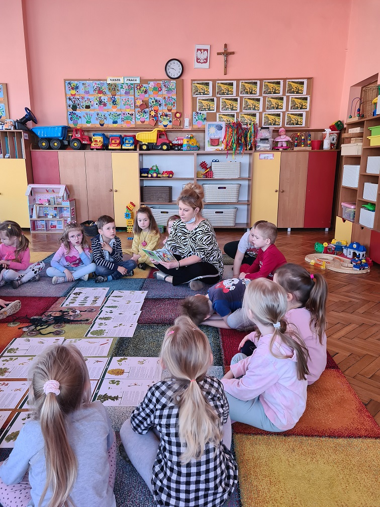 Dzieci siedzą w kole po "turecku" i słuchają czytanego przez nauczycielkę opowiadania.