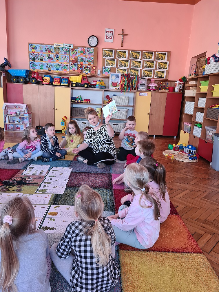 Nauczycielka pokazuje dzieciom na obrazku leśniczego - gospodarza lasu. Dzieci siedzą w kole po "turecku".