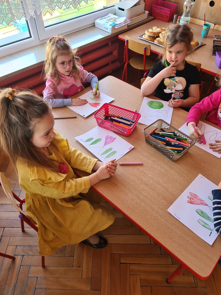 Dzieci przy stoliku kolorują kredkami swoje rysunki.