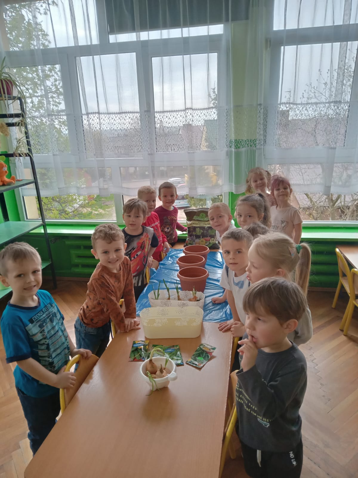Grupa dzieci przy stolikach przygotowuje się do sadzenia nasion w doniczkach z ziemią