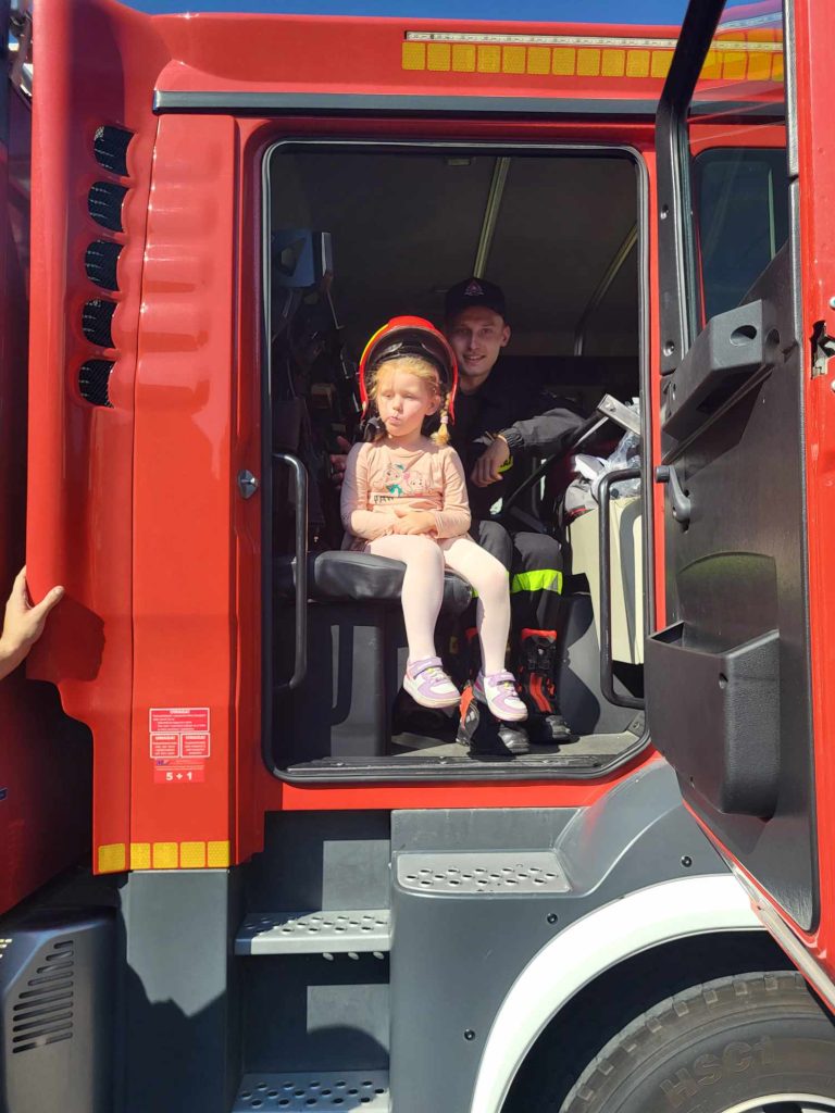 Dziewczynka w hełmie strażackim siedzi w wozie strażackim