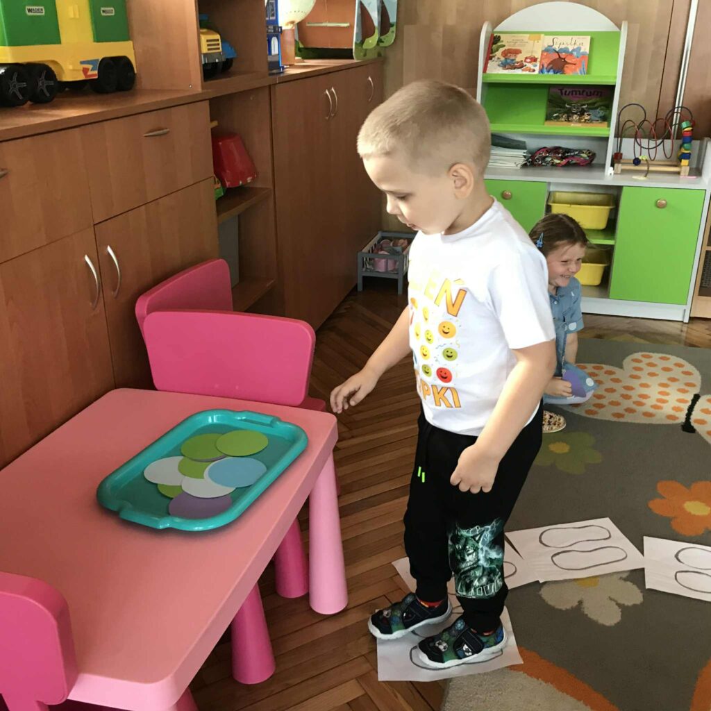 Dzieci siedzą na dywanie, chłopiec idzie po kartkach z narysowanymi śladami stóp.