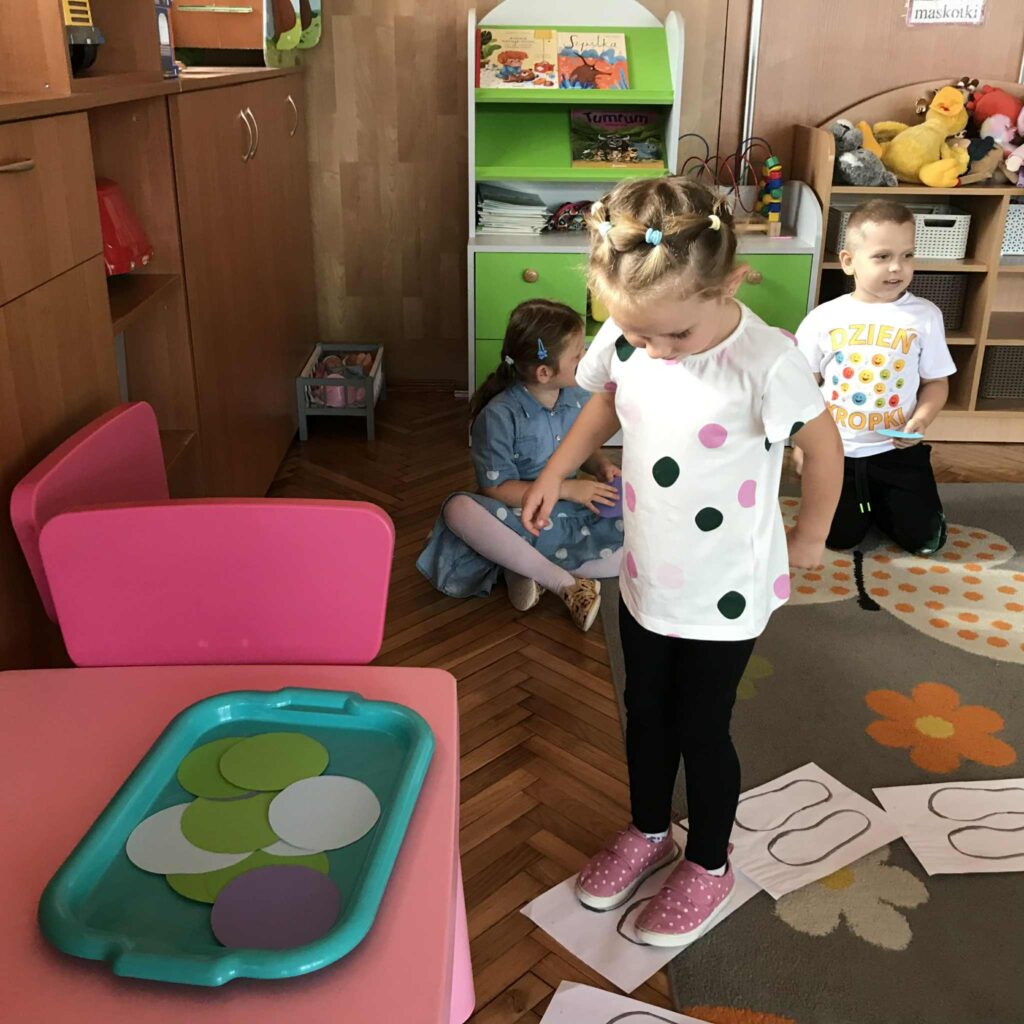 Dzieci siedzą na dywanie, dziewczynka idzie po kartkach z narysowanymi śladami stóp.