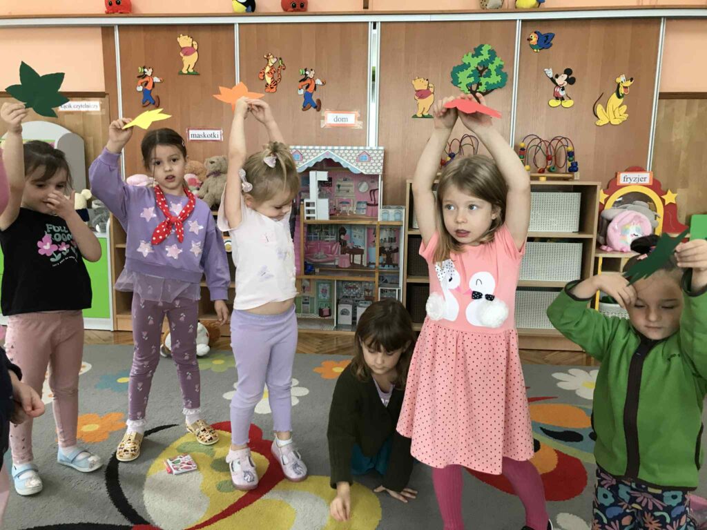 Dzieci stojąc zrzucają na dywan papierowe liście.